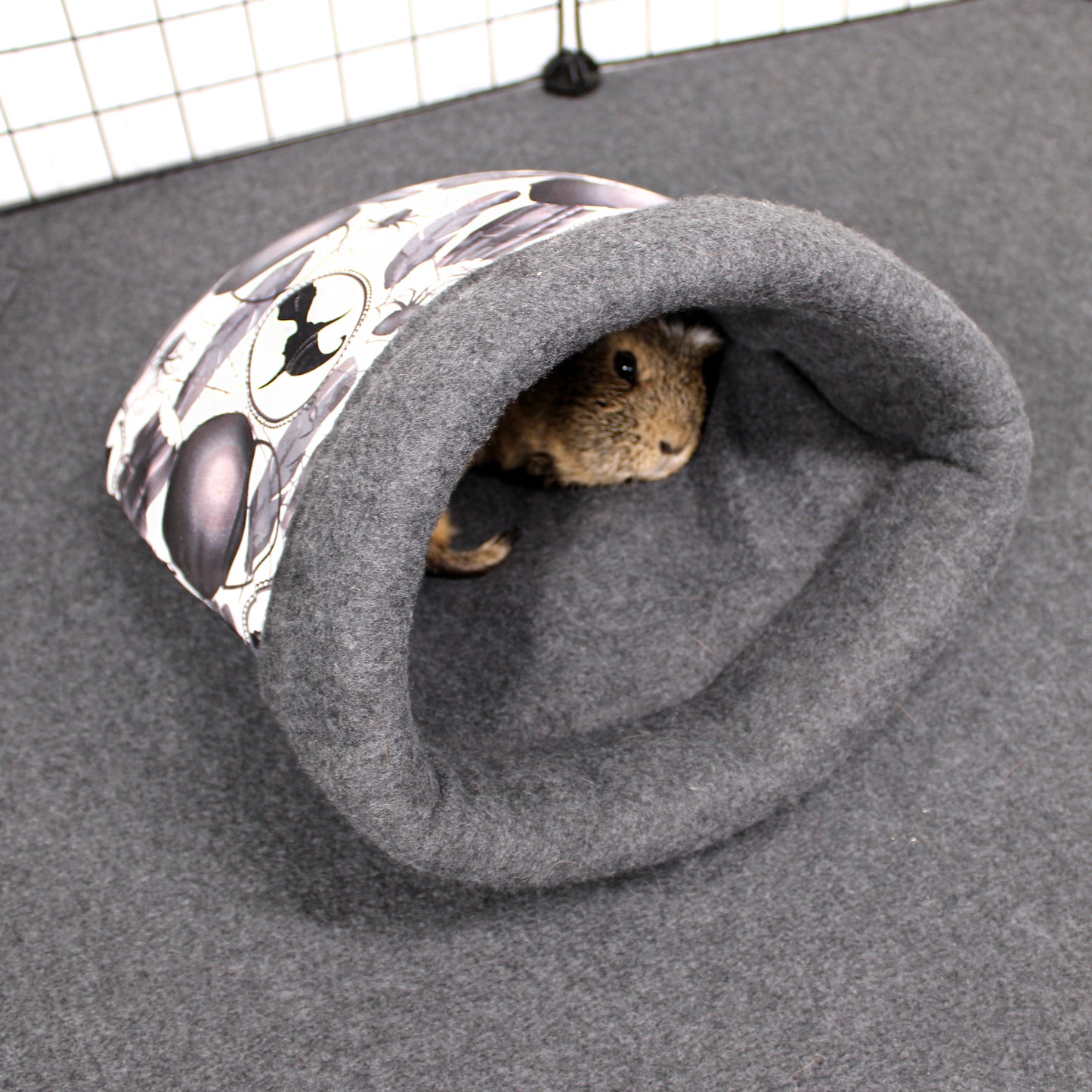 Small animal peeking out from a gray cuddle sack with a patterned entrance on a gray surface.