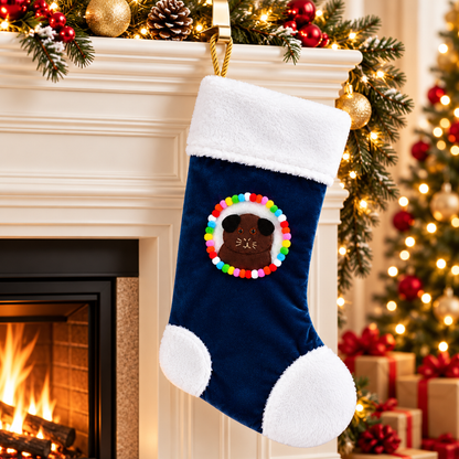 Chocolate brown guinea pig Christmas stocking with darker brown ears hanging beside a decorated Christmas fireplace, featuring a handmade felt guinea pig character, navy blue velvet, fluffy white trim, colourful pom-poms and gold rope hanging loop.