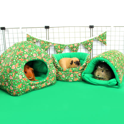 Three guinea pigs in green gingerbread pet houses on a green surface with a wire cage background.